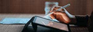 A close-up of a person's hand using a stylus to write or draw on a tablet, set on a wooden desk with papers in the background, shot in warm, moody lighting.
